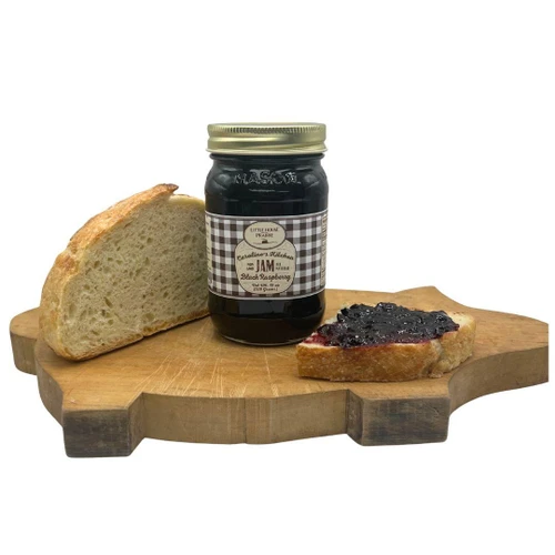 A jar of Caroline's Kitchen Black Raspberry Jam sits on a uniquely shaped pig wooden cutting board next to a slice of crusty bread and a slice spread with dark jam, displayed against a solid white background.