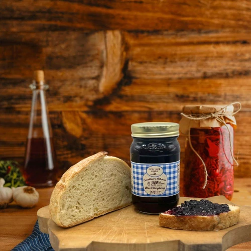 A jar of Caroline's Kitchen Black Raspberry Jam sits on a wooden cutting board next to a slice of fresh crusty bread and a slice spread with dark jam, set against a rustic wooden background.
