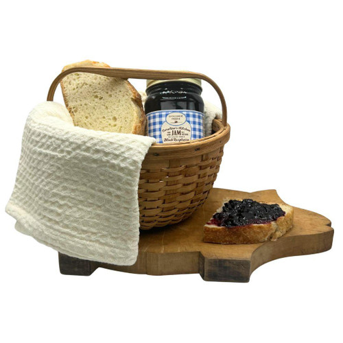 A jar of Caroline's Kitchen Black Raspberry Jam nestled in a woven basket next to fresh bread, sitting on a uniquely shaped pig wooden cutting board with a slice spread with dark jam, displayed against a solid white background.