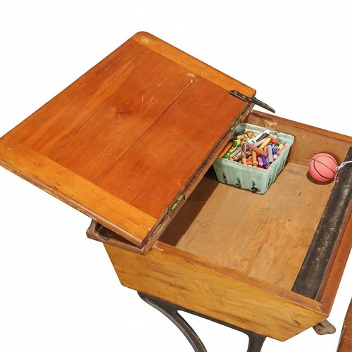 The interior storage compartment of the vintage school desk is shown open, revealing a wooden storage space. Inside, there is a green plastic basket filled with crayons alongside a small pink toy basketball. The hinged wooden desktop is propped open to the left, highlighting its grain and metal hardware against a plain white backgroun