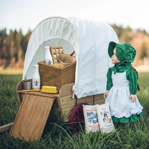 A pioneer doll in a green dress and bonnet standing next to a miniature covered wagon filled with vintage baking supplies in a grassy field.