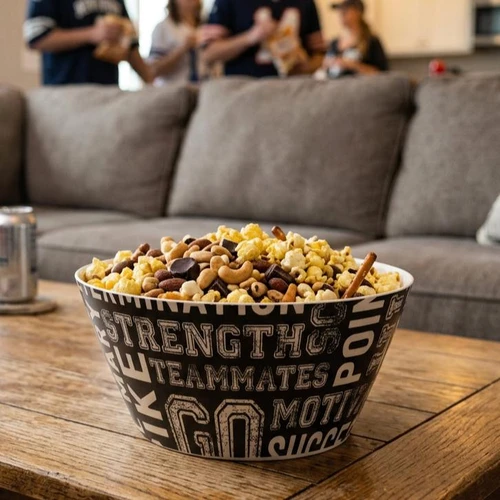 A black serving bowl printed with motivational sports words like "STRENGTH" and "TEAMMATES" filled with a snack mix sits on a rustic wooden coffee table. The mix includes popcorn, cashews, almonds, chocolate pieces, and pretzel sticks, while a blurred grey couch and people in sports jerseys appear in the background.