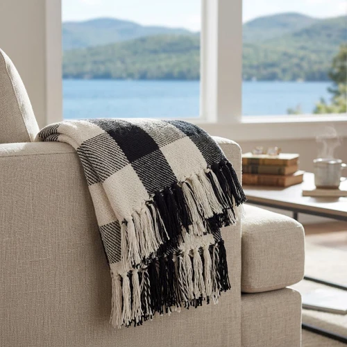 A black and white buffalo check plaid woven throw blanket with thick fringed edges is draped over the armrest of a beige fabric armchair. In the background, a large window reveals a serene lake and mountain landscape.