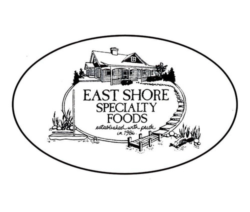 An oval-shaped black and white logo for East Shore Specialty Foods. The center features a detailed line-art illustration of a rustic lakeside house with a smoking chimney. Below the house, a sign reads "EAST SHORE SPECIALTY FOODS established with pride in 1986." A small wooden pier and a canoe rest in the water in the foreground, surrounded by reeds and rocks.