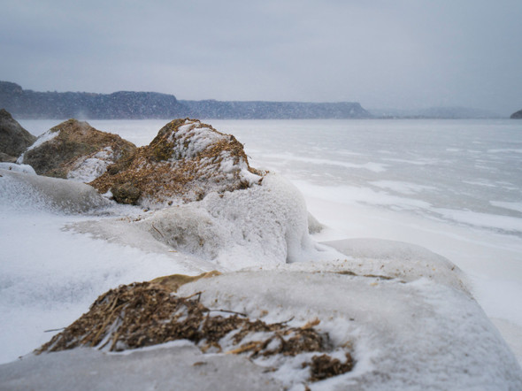 ice covered rocks jutting out into an icy lake with mountains in the distance and snow flurries in evidence