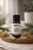 Essential7 Happy Bones essential oil blend in an amber glass bottle with the original Essential7 logo, displayed on a natural wooden tray and surrounded by botanicals that reflect the blend’s ingredients. White Spruce and Balsam Fir branches frame the bottle, while Helichrysum flowers, Lemongrass, Vetiver root, and Copaiba resin create a fresh, earthy presentation. Soft natural lighting, neutral linen, and a gently blurred background give the image a warm, grounded, forest-inspired feel.