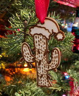 A wooden ornament in the shape of a saguaro cactus wearing a Santa Claus style hat with christmas lights around it. Hung with a red ribbon in a fir tree
