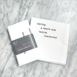 a marble countertop with a white towel laying on top on it, folded in half with the text in a typewriter font. For the full text please see product description. Next to the towel is the folded version of the same towel with a gray band around it with the Dev D and Co. logo