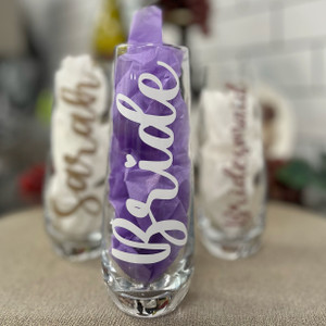 Clear glass stemless champagne flute, with purple tissue paper on the inside. The glass reads "Bride" in white cursive font and a heart acting as the dot for the "i"
