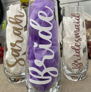 Three stemless champagne flutes, two behind and one in front. The two in the back read "Sarah" and "Bridesmaid". The one in front reads "Bride". All in cursive font horizontally down the glass