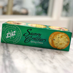 Rectangular green box with the photo of a cracker on it; a round yellow cracker with flakes of green in it. The Elki logo and wording "Savory Crackers Spring Onion" are on the top side of the box