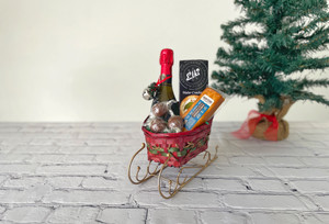 Wide shot of a red and green sleigh basket with green filling. There is a wine bottle, crackers, cheese and chocolates in it. There is a green Christmas tree in the background. Please see description for product details. 