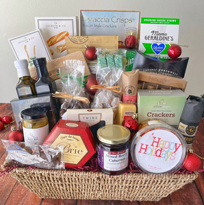 A woven brown basket with burgundy filling and products standing in it. Red ornaments are placed in and around the basket.