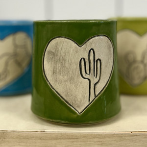 A dark green drinking glass with a heart, inside of the heart is off-white in color depicting a saguaro cactus