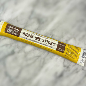 marble countertop with a rectangular stick diagonal in the middle. The yellow packaging reads the name of the company, "Singing Pastures" as well as the title of the product, "Roam Sticks" with an icon of a pig in between the words