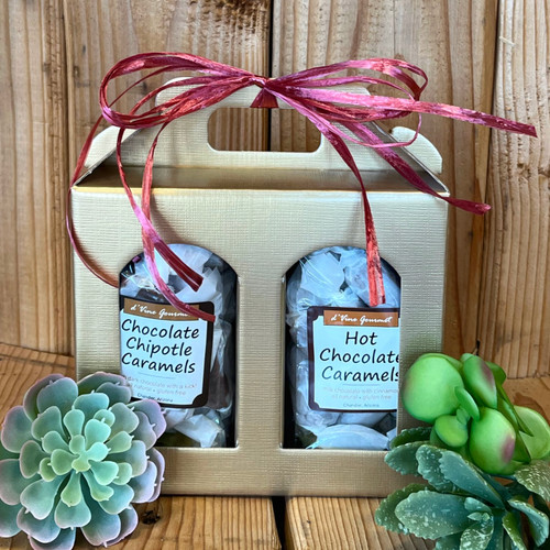 A linen patterned gold cardboard box with two arched windows on the front. Each window has a bag of caramels showing: one with Hot Chocolate Caramels, and one with Chocolate Chipotle Caramels. Box is tied with a burgundy raffia bow 