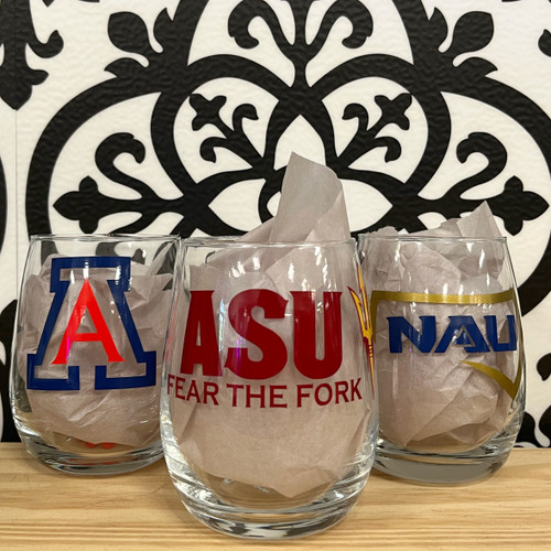 Three Clear stemless wine glasses displayed on a wood table of the Arizona universities' logos