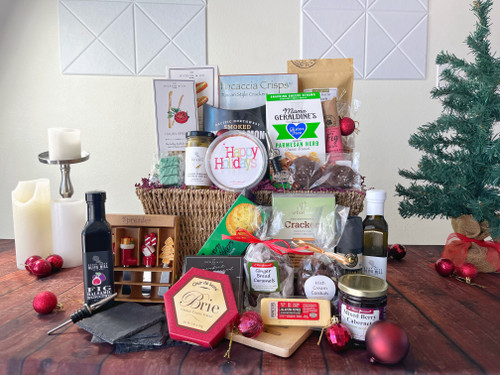 A woven brown basket with products standing in it and on the table in front of it. Three candles and a green Christmas tree are in the background. Red ornaments are in and around the basket.