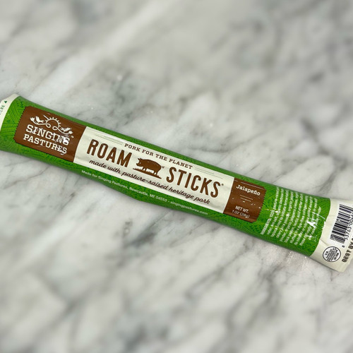 marble countertop with a rectangular stick diagonal in the middle. The green packaging reads the name of the company, "Singing Pastures" as well as the title of the product, "Roam Sticks" with an icon of a pig in between the words