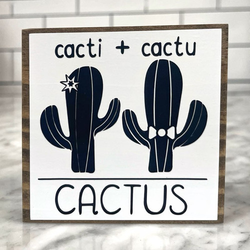 Square wood block with white background with black lettering and a black cactus with a star and another black cactus with a bowtie.