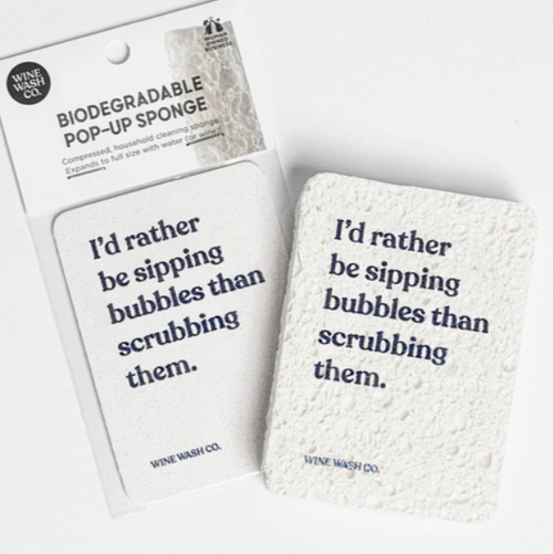 White rectangular sponge on white cardboard display packaging and a second open sponge with black lettering that reads "I'd rather be sipping bubbles than scrubbing them"