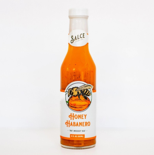 Tall clear glass bottle showing orange colored contents with white label and orange lettering and image of a bee on a habanero pepper 