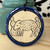 circular ceramic ornament with a dark blue border around an off-white circle depicting a javelina next to two prickly pear cacti
