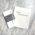 a marble countertop with a white towel laying on top on it, folded in half reading " ' Alexa ... pour more wine' " in a typewriter font. Next to the towel is the folded version of the same towel with a gray band around it with the Dev D and Co. logo