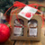 A linen patterned gold cardboard box with two arched windows on the front. Each window has a bag of caramels showing: one with Classic Sea Salt Caramels, and one with Gingerbread Caramels. Box is tied with a red raffia bow and a tag reading "Happy Holidays"