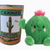 Plush barrel cactus with arms, legs, eyes and pink puff of hair next to a yellow and green can displaying the front label with black lettering