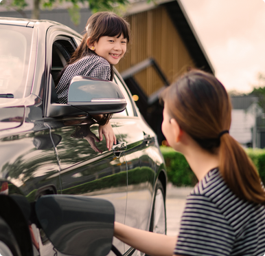 A child leans out of a car window, smiling at their parent who is plugging in a home EV charger.