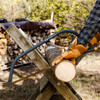 A gloved hand steadying a log while cutting it with the Heavy Duty Bow Saw beside a woodpile