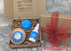 3-piece shampoo & sea salt hair spray gift set — Salty Mermaid: the image depicts a silver background on which is placed two gift boxes. One is open and displaying its contents; the other is still closed and sealed with a red bow. The open box contains — nestled in a bed of crinkle paper — a bright blue shampoo bar, a copper shampoo bar carrier with a blue label on the lid, and a small plastic bottle of sea salt hair spray with a label. All three items are labeled "Salty Mermaid" scent.
