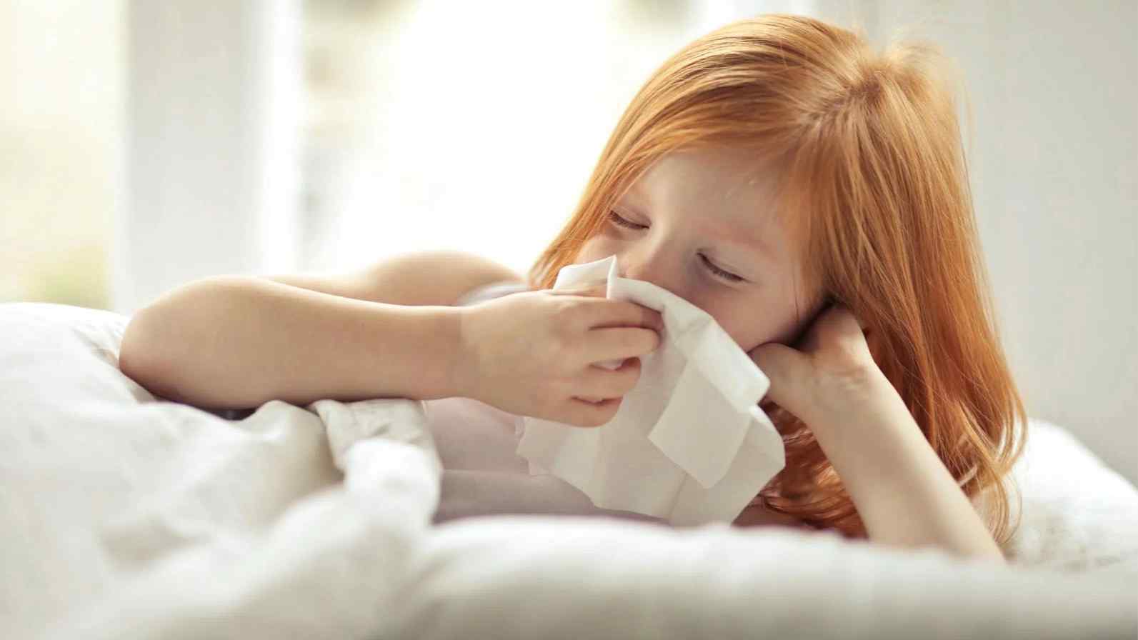A young girl with red hair uses a tissue to blow her nose, illustrating the impact of allergies and hay fever in children.