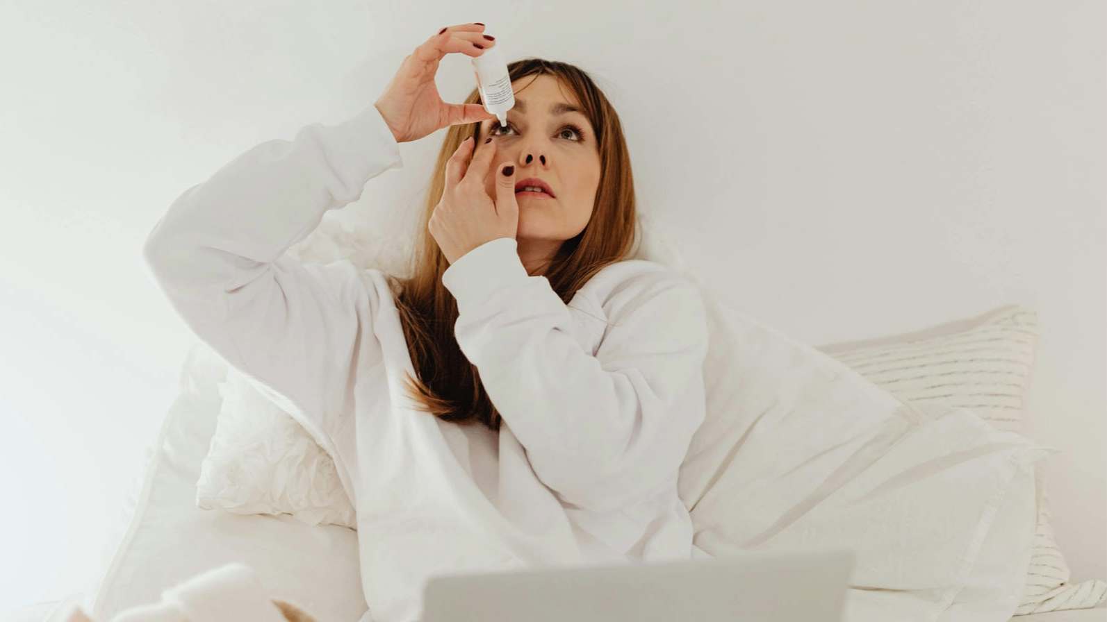 A woman sitting on a bed, using a laptop while applying eyedrops to her eyes.