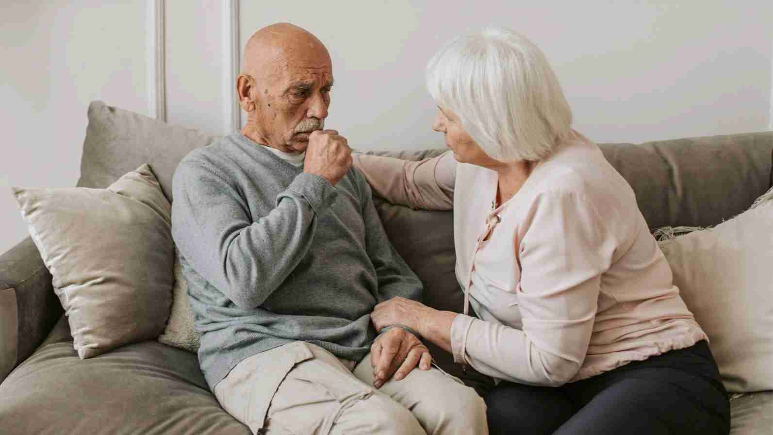 An older man with a chesty cough and a woman sitting together on a couch, sharing a moment of companionship.