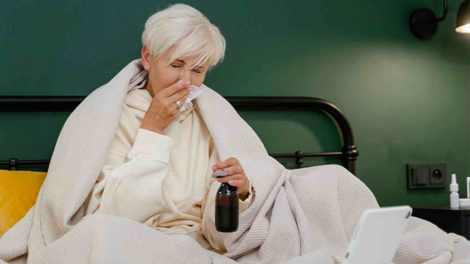 An older woman in bed with a laptop, showing signs of illness, likely experiencing cold or flu symptoms.