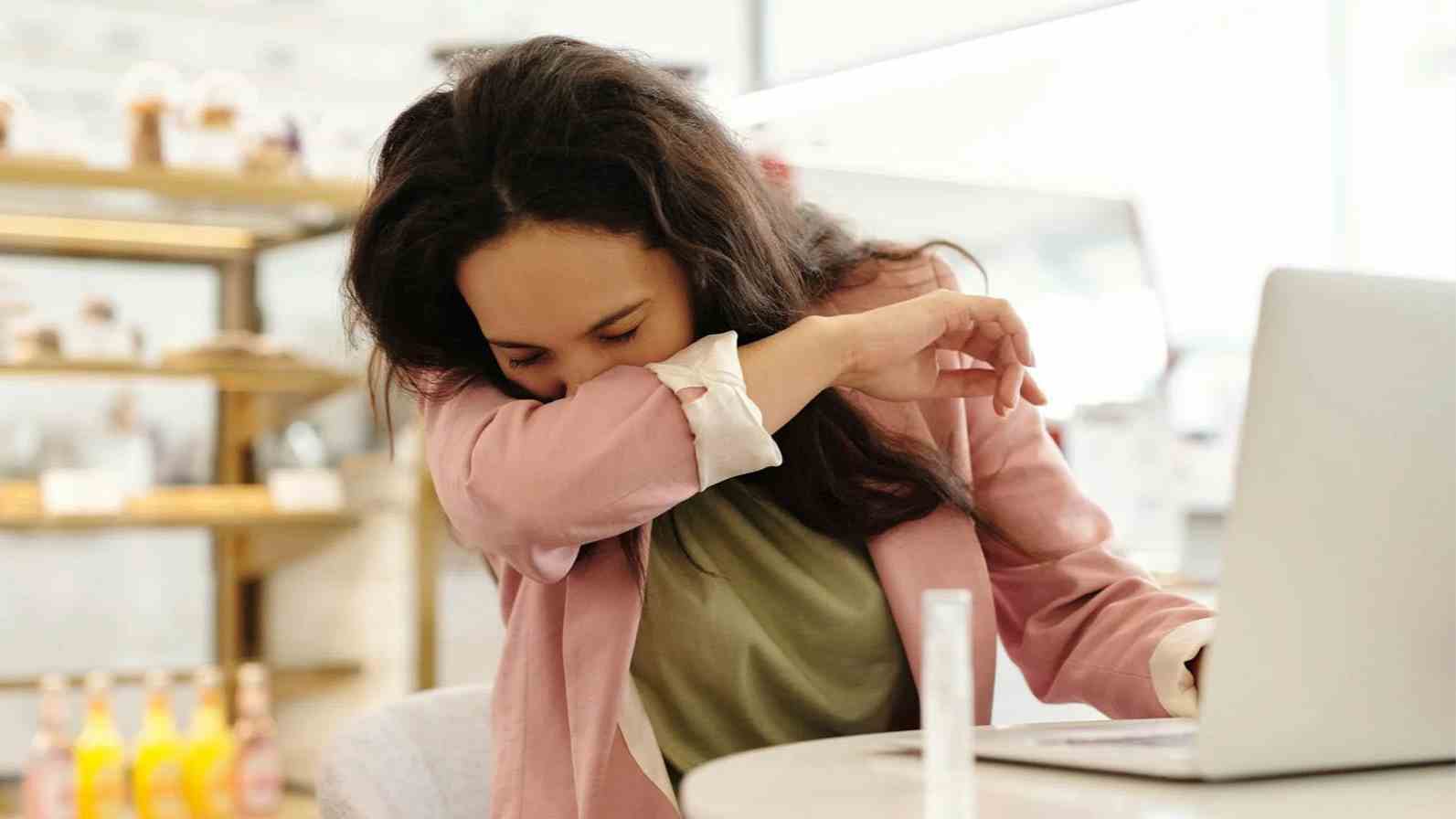 A woman at a table with her laptop, appearing to sneeze, possibly due to histamine allergies.