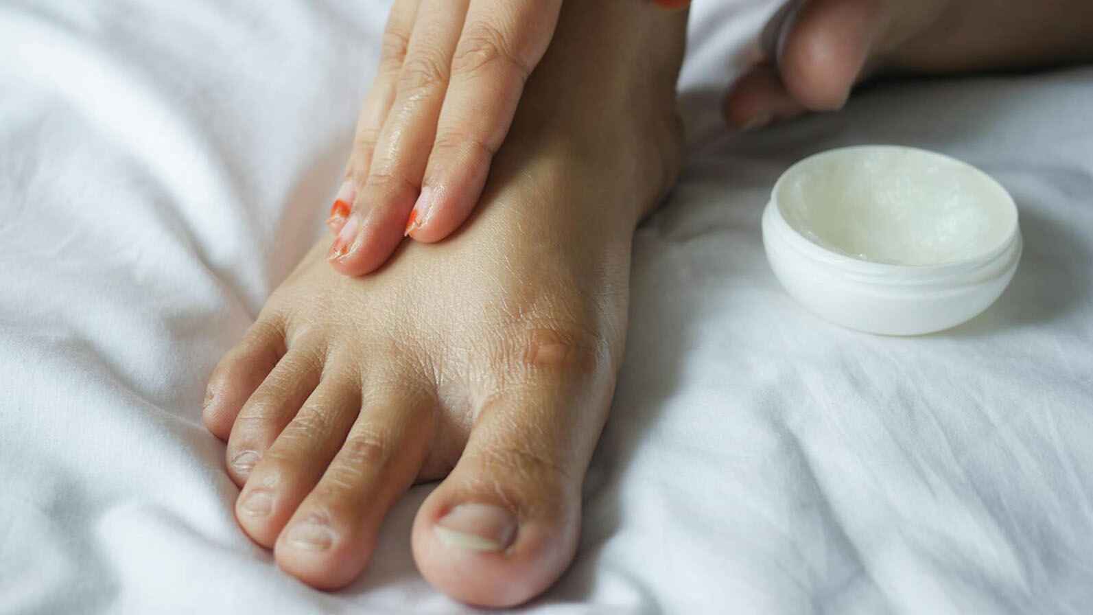 A person's feet resting on a bed beside a jar of cream, highlighting the connection between hands and feet in self-care.