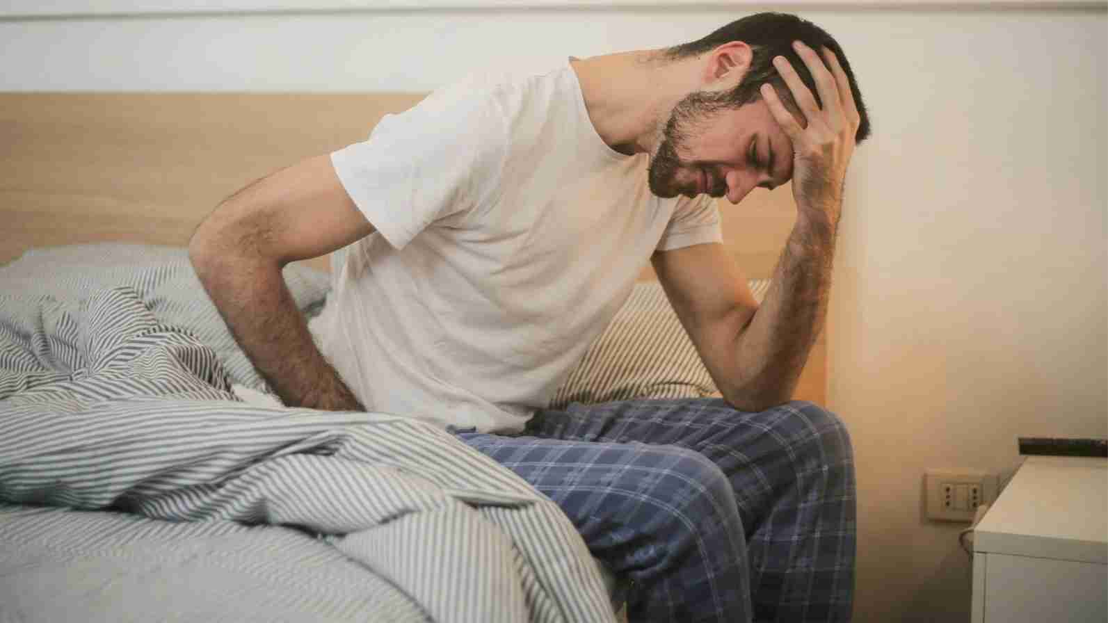 A man sits on his bed, head in hands, conveying the distress of a headache while searching for pain relief.