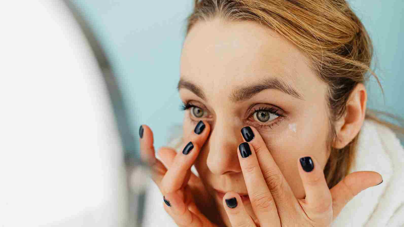 A woman applies eye cream while gazing into a mirror.