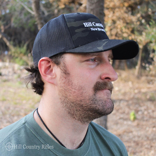 HCR Multi Cam Trucker Hat, black and gray camouflage patterned mesh back with black front panel and bill, logo embroidered on front, worn by a man outdoors.