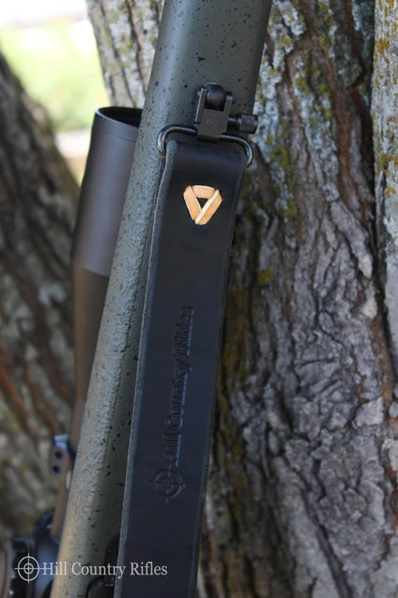 HCR Murray Leather HCR Rifle Sling in Black, close-up of the padded leather strap with embossed Hill Country Rifles logo and metal swivel hook, attached to a rifle against a tree bark background.