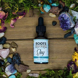 1 oz dropper bottle of Roots to Remedies Joy Herbal Extract displayed on a wooden background surrounded by colorful crystals, flowers, and moss.