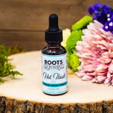 A 1 oz amber glass dropper bottle labeled “Hot Flash” by Roots to Remedies is positioned upright on a natural wooden slab. The label is light blue with black and white text that reads “Menopause Support Formula – Alcohol Free.” The bottle is surrounded by fresh flowers, including a pale pink dahlia, purple statice, and green mums, creating a soft and supportive botanical setting that reinforces the product’s natural and feminine purpose.