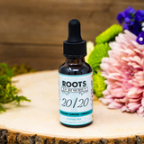 An amber glass dropper bottle labeled "Roots to Remedies – 20/20 Eyesight Support Formula" stands on a wooden surface, surrounded by colorful flowers including pink chrysanthemums, green moss, and purple blossoms. The label notes that the formula is alcohol-free.