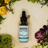 A small amber glass dropper bottle labeled "Roots to Remedies – Balancing Act" sits on a light wood surface surrounded by dried herbs and flowers, including moss, rose petals, and yellow blossoms. The label indicates it is an alcohol-free hormone balancing formula.