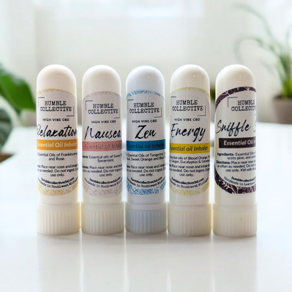 A lineup of five white essential oil inhalers from Humble Collective, each labeled with a different blend: Relaxation, Nausea, Zen, Energy, and Sniffle. The inhalers are standing upright on a white surface with blurred green plants in the background, showcasing clean and minimal packaging design. A lineup of five white essential oil inhalers from Humble Collective, each labeled with a different blend: Relaxation, Nausea, Zen, Energy, and Sniffle. The inhalers are standing upright on a white surface with blurred green plants in the background, showcasing clean and minimal packaging design.