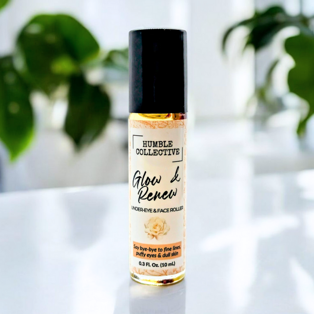 A 10 mL glass roller bottle of Glow & Renew Under-Eye & Face Roller by Humble Collective is displayed upright on a glossy white surface with lush green plants softly blurred in the background. The label reads “Glow & Renew – Under-Eye & Face Roller” in elegant script, with a golden accent. A small green badge in the corner reads “THC FREE.” The overall aesthetic is clean, bright, and natural—highlighting the product’s purity and skincare benefits.