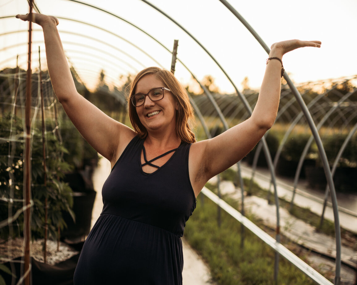 Sarah, the founder of Humble Collective, stands joyfully with arms raised inside a greenhouse tunnel at sunset. She’s wearing a navy sleeveless dress and glasses, surrounded by rows of healthy hemp plants. The golden light and thriving greenery reflect the heart and energy behind her wellness brand.